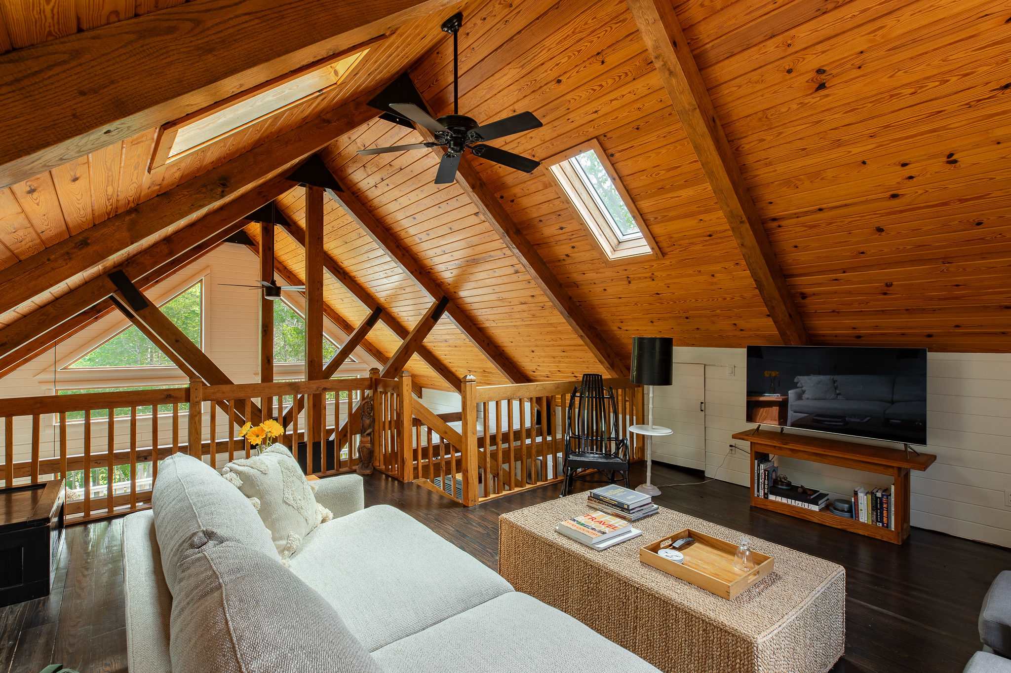 A cozy loft area serves as a secondary living space, featuring a comfortable sectional sofa and a TV on a low media console. The space is defined by its warm wooden vaulted ceiling with skylights and exposed beams, overlooking the main living area below.