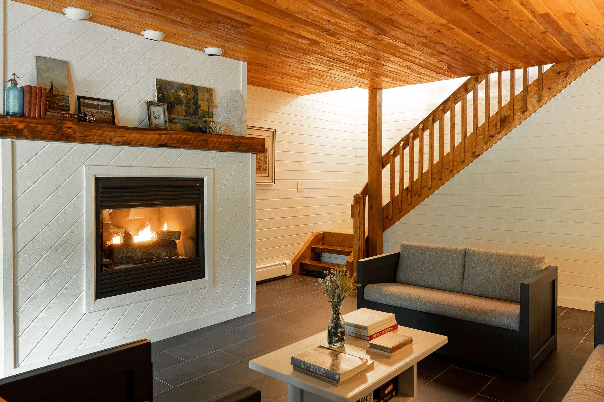 Inviting living room with a modern fireplace set into white shiplap walls, featuring a grey sofa and a light coffee table. A wooden staircase with open risers leads to an upper level, and the ceiling is also wood-paneled.