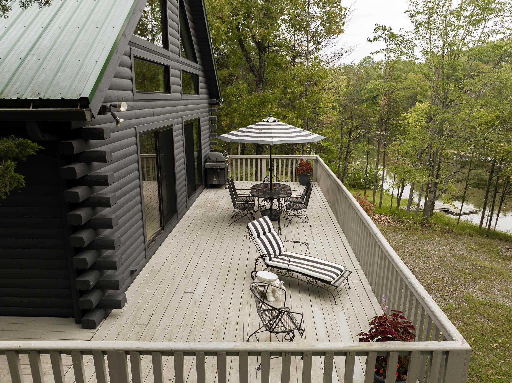 Spacious outdoor deck with a black and white striped umbrella shading a dining table and chairs, along with lounge chairs, all overlooking a serene lake. A dark log cabin exterior with a green roof is visible.