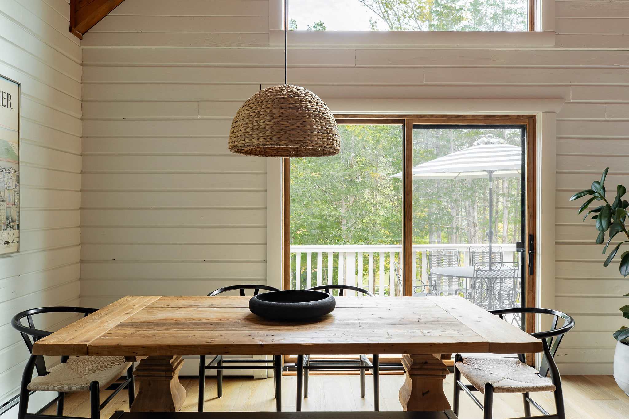 Rustic dining area with a large wooden table, black chairs, and a woven pendant light, featuring white shiplap walls and a sliding glass door leading to a deck with lake views.