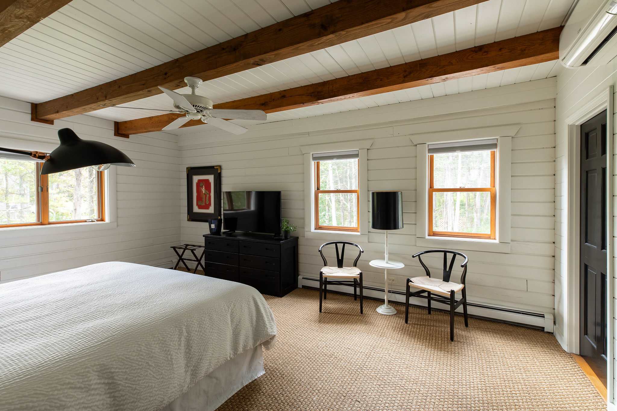 Bright bedroom featuring white shiplap walls, exposed wooden ceiling beams, and a ceiling fan. A large bed with white bedding faces a black dresser with a TV, flanked by two windows and a seating area.