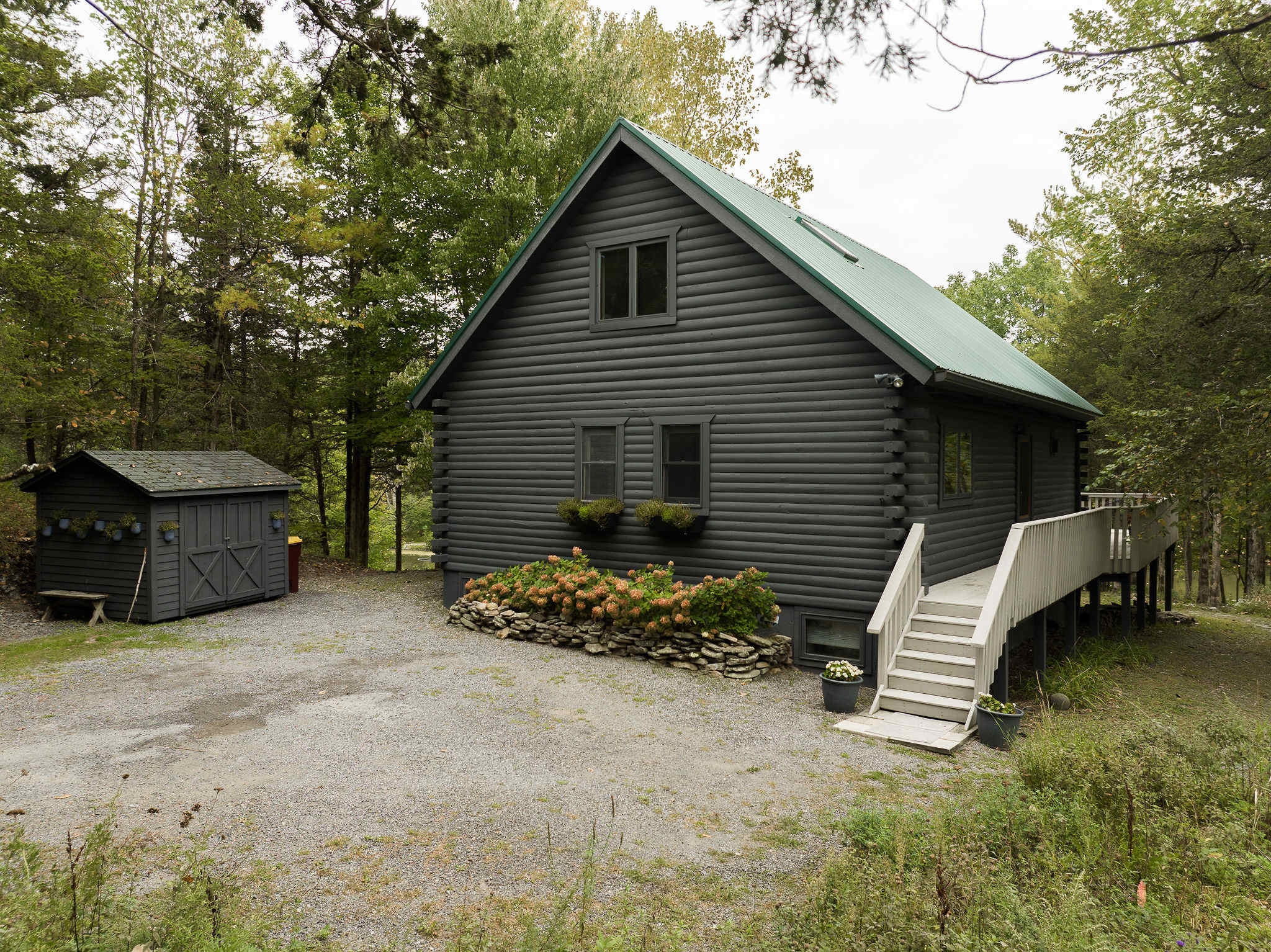 A side view of the gray log cabin house, featuring a green metal roof, a small shed, and white stairs leading up to a front entrance and deck, surrounded by lush greenery.