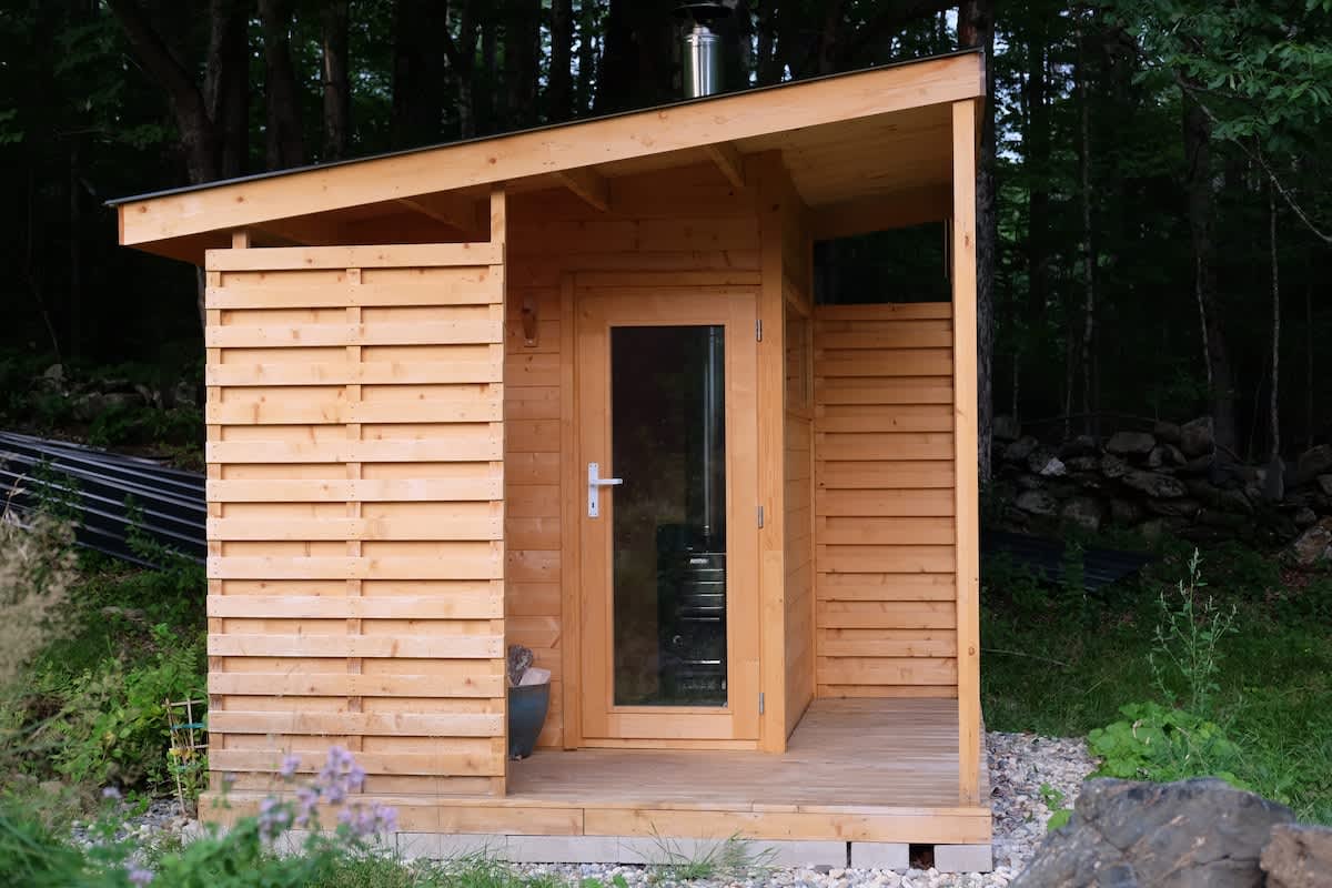 Exterior view of a modern wooden sauna building with a glass-paneled door, featuring a sloped roof and slatted sides, nestled among trees and greenery.