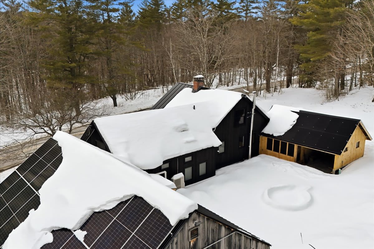 Aerial view of a black farmhouse and outbuildings covered in snow, with solar panels visible on one roof. The property is surrounded by a mix of bare and evergreen trees in a winter setting.