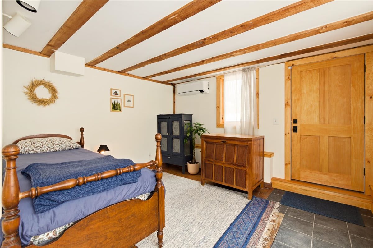 View of a rustic bedroom with a wooden single bed, exposed ceiling beams, and a mix of slate and wood flooring. A wooden door and a small window are visible, along with a dark cabinet and a light wood dresser.