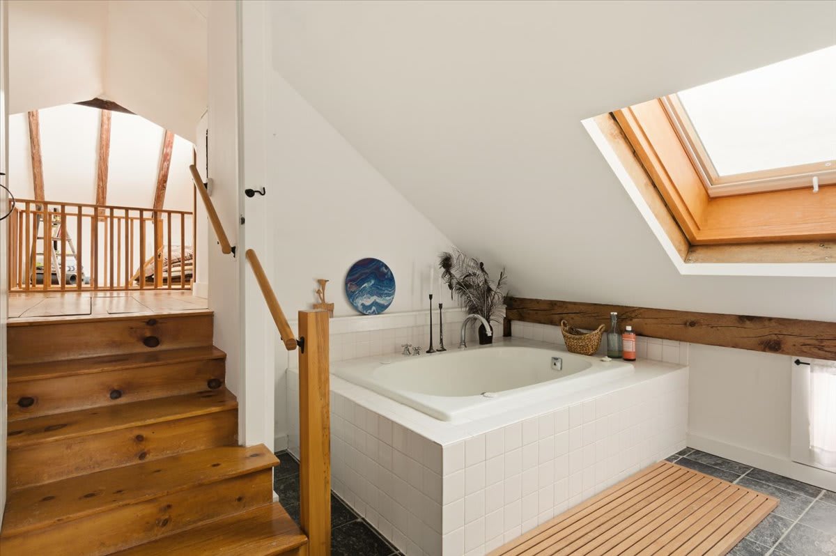 An attic bathroom features a built-in white tiled bathtub under a skylight, with wooden stairs and railing leading up to another level. Exposed wooden beams add a rustic touch to the bright space.