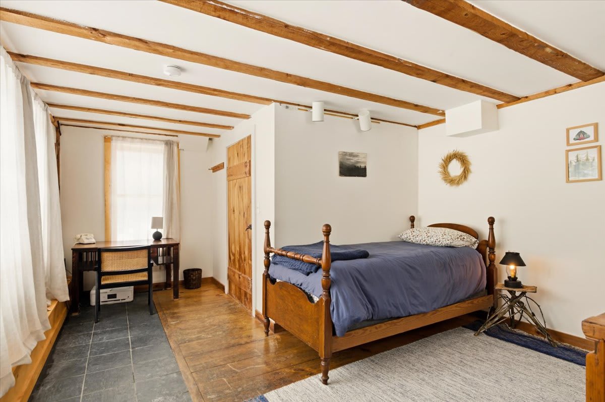 A cozy bedroom featuring a wooden single bed with blue bedding, exposed wooden ceiling beams, and a desk area with a window. The floor transitions from dark slate tiles to light wood planks.