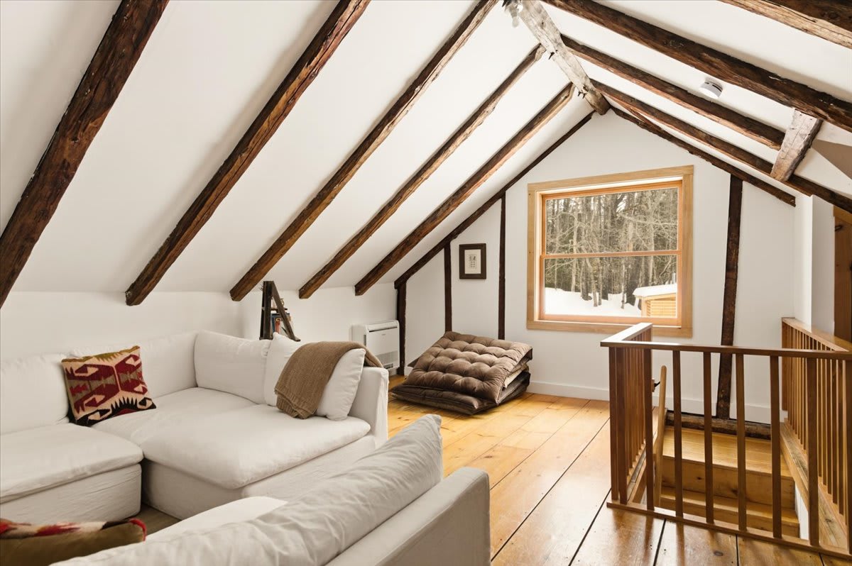 A bright living area in a loft space showcases white slipcovered sofas, a brown floor cushion, and a large window offering views of a snowy forest, all under a vaulted ceiling with exposed wooden beams. A wooden railing indicates a staircase.