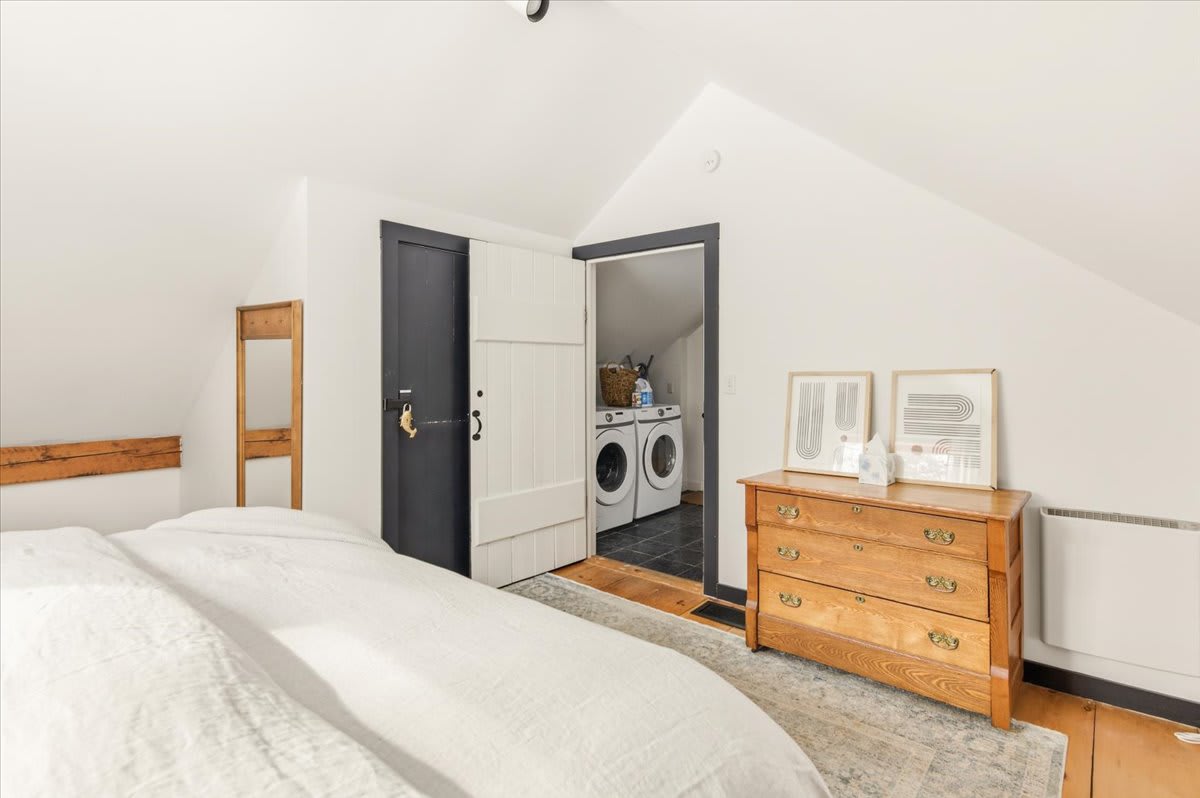 View of the attic bedroom showing a wooden dresser and a dark barn-style door leading to an adjacent laundry area. The room has a sloped ceiling, light hardwood floors, and a rustic exposed beam.