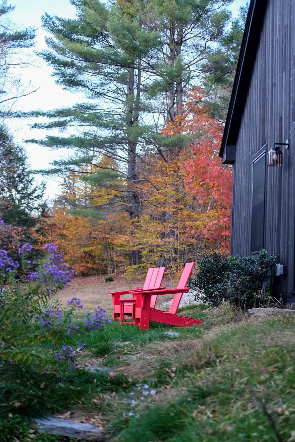 Two vibrant red Adirondack chairs sit on a grassy slope beside the dark-sided house, overlooking a field of purple flowers and a backdrop of stunning autumn foliage.