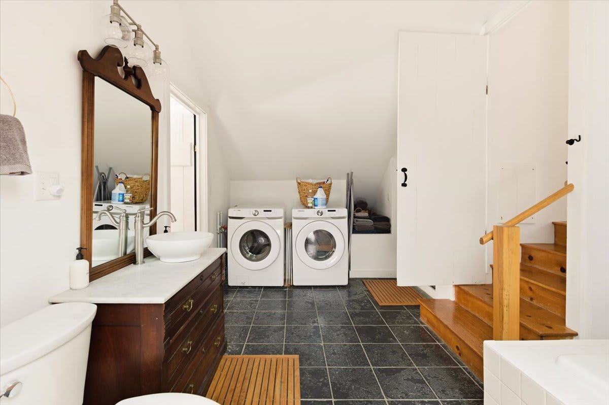 A spacious attic bathroom featuring a dark wood vanity with a vessel sink, a toilet, and two white washing machines. Dark slate floor tiles and white walls create a clean, functional space with wooden stairs leading to another level.