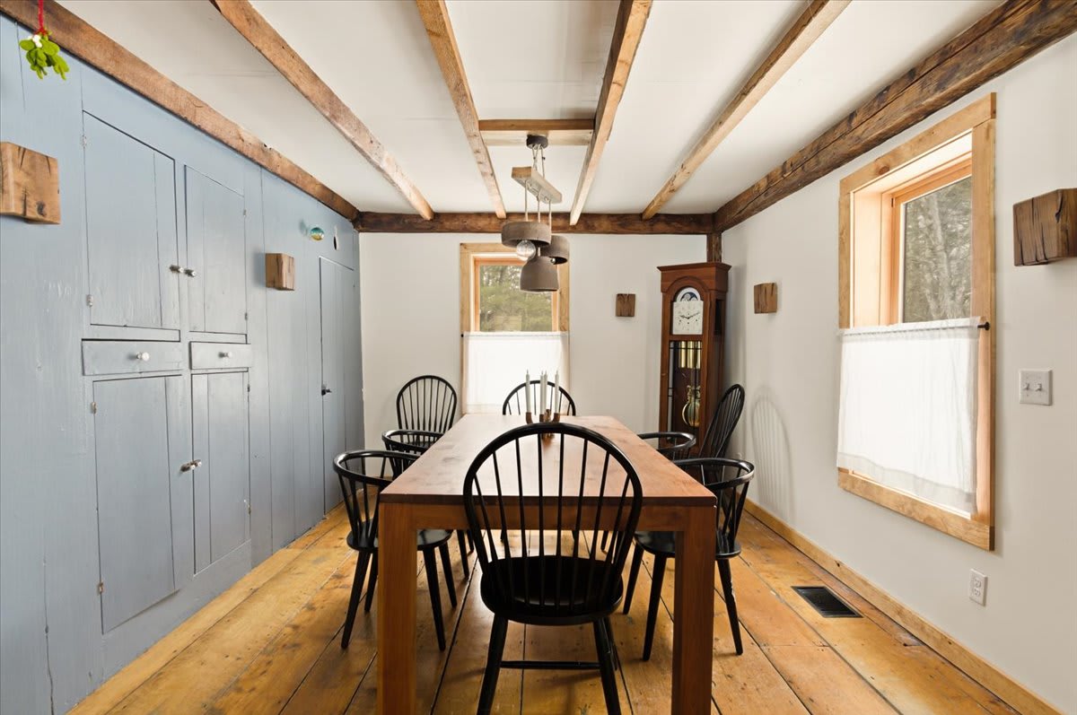 A rustic dining room features a long wooden table with six black spindle-back chairs, set against a wall with light blue built-in cabinets and exposed wooden ceiling beams. A tall grandfather clock stands in the corner.