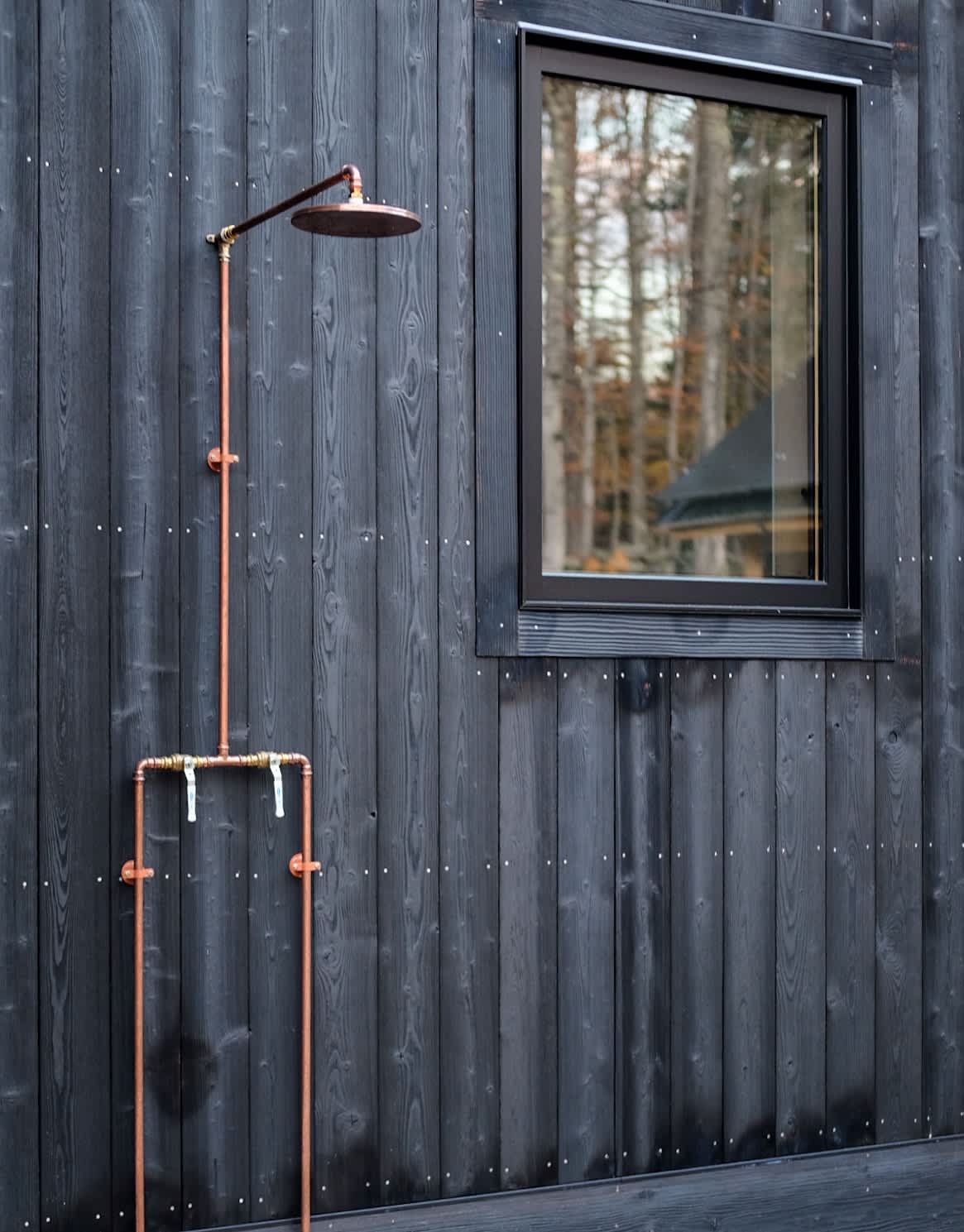 An outdoor shower setup with exposed copper piping and a rain shower head, mounted on the dark vertical siding of the house, adjacent to a large black-framed window.