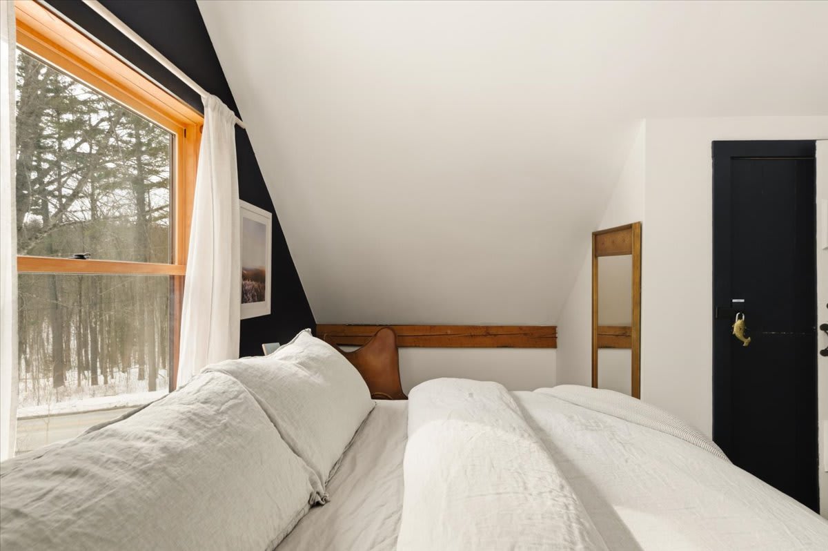 Close-up of the bed in the attic bedroom, showing light bedding and a large window with wooden trim offering views of a snowy forest. A dark accent wall is visible behind the bed, complementing the white sloped ceiling.