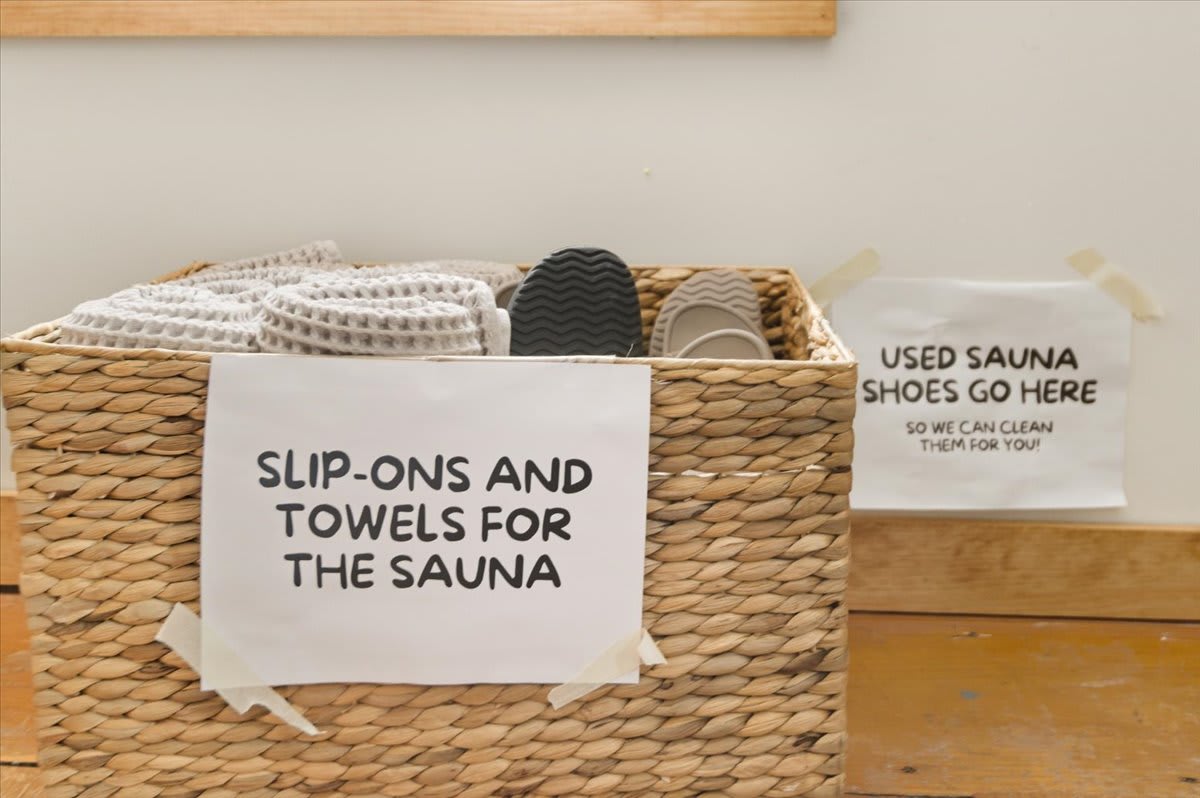 A wicker basket holds white waffle-knit towels and dark slip-on shoes, with signs indicating "SLIP-ONS AND TOWELS FOR THE SAUNA" and a collection point for "USED SAUNA SHOES SO WE CAN CLEAN THEM FOR YOU!".