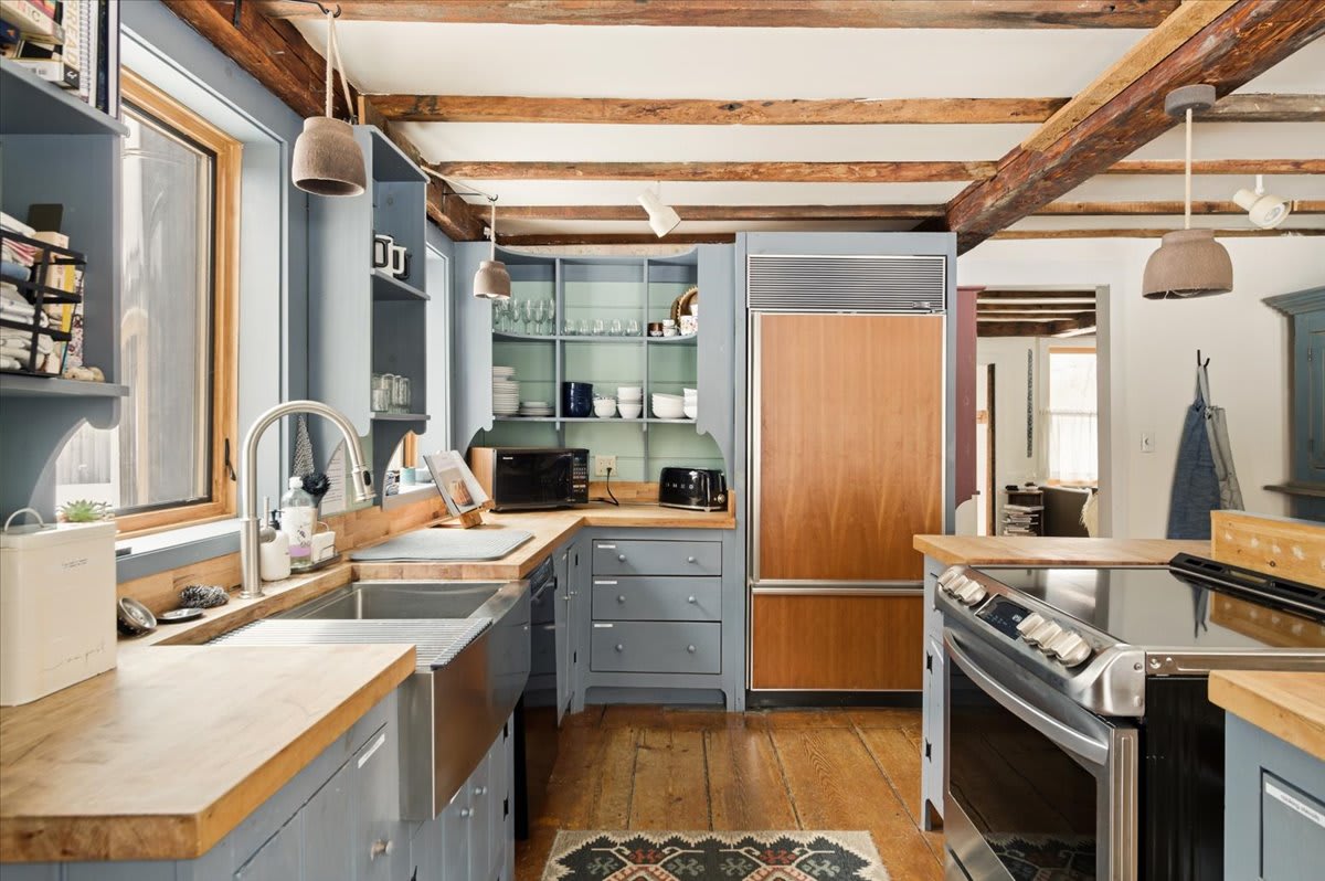 A charming kitchen with light blue cabinetry, butcher block countertops, and exposed wooden beams. It features a stainless steel oven, a wood-paneled refrigerator, and rustic hardwood floors.