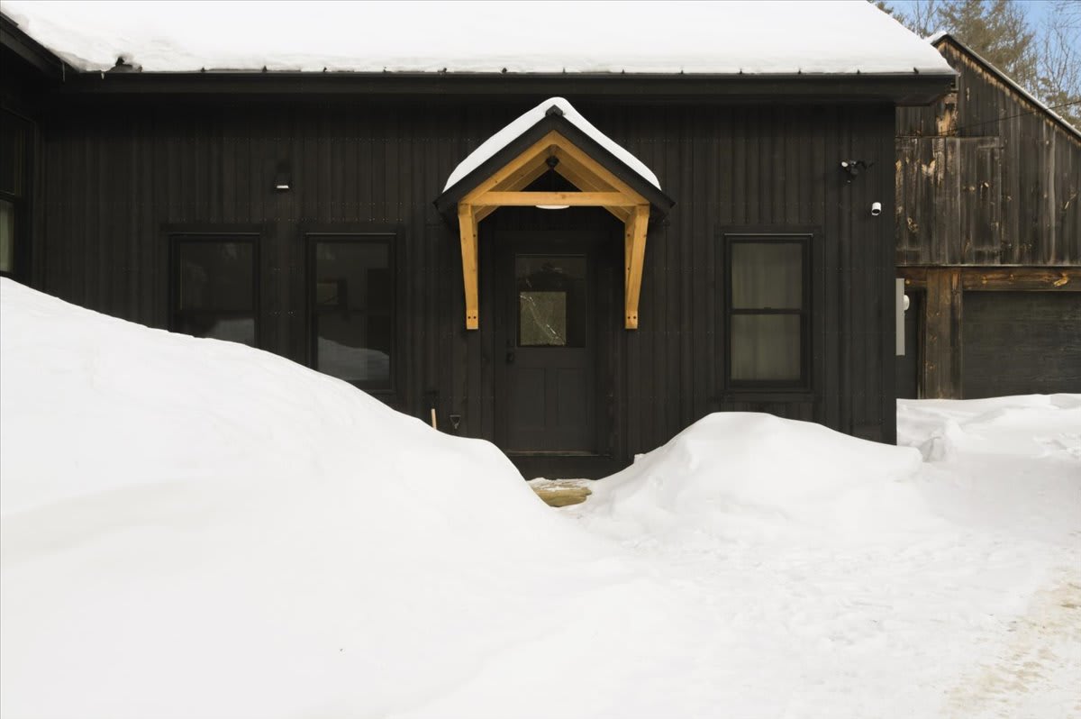 Front view of a dark-colored farmhouse with a wooden-framed entrance canopy, surrounded by deep snowdrifts, under a bright winter sky, highlighting the main entry point.