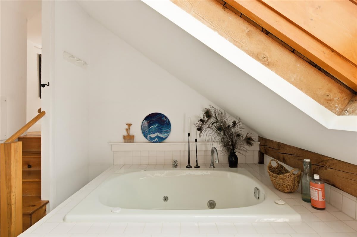 Close-up of the white built-in bathtub in the attic bathroom, surrounded by white tiles and featuring a skylight above. Decorative items and exposed wooden beams enhance the rustic aesthetic.
