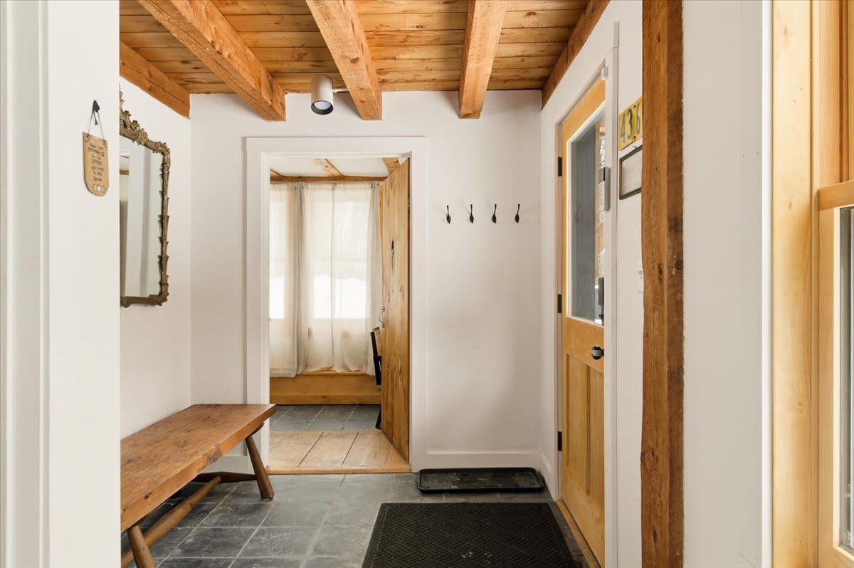 Bright entryway with dark slate tile floors, exposed wooden ceiling beams, a rustic wooden bench, and a decorative mirror. A wooden door with a glass panel leads outside.