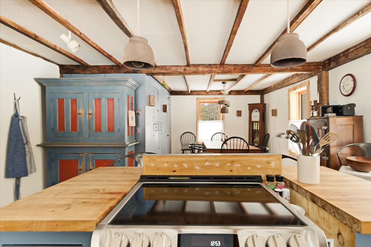 A spacious open-concept room featuring a dining area with a rustic table and a striking blue and red painted cabinet. Exposed wooden beams crisscross the ceiling, complementing the hardwood floors and farmhouse aesthetic.