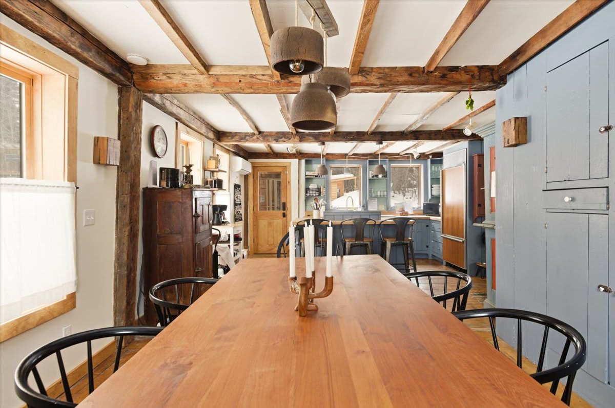 The dining room, with its long wooden table and black chairs, opens into a rustic kitchen featuring light blue cabinetry, a breakfast bar with stools, and exposed wooden ceiling beams throughout.