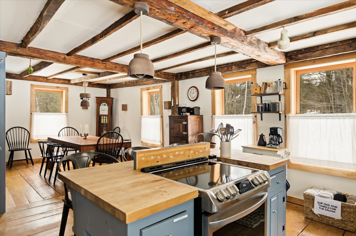 An inviting open-concept dining area with a rustic wooden table and black chairs, set against a backdrop of exposed wooden beams and hardwood floors. The kitchen island with a stovetop is visible in the foreground.