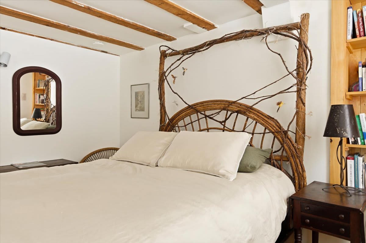 View of a queen bed with a natural branch-style headboard and frame, set against a white wall with exposed ceiling beams. A small mirror and artwork are visible, reflecting the rustic charm of the room.