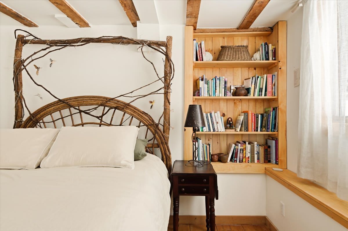Close-up of a queen bed with a distinctive branch-style headboard, flanked by a small wooden nightstand with a lamp and a built-in wooden bookshelf filled with books. Exposed ceiling beams and hardwood floors are visible.