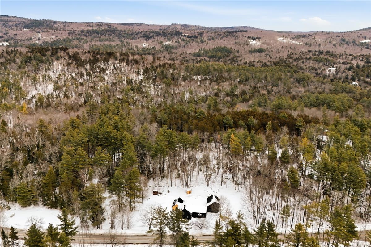 Expansive aerial view showcasing the property's dark buildings amidst a sprawling, snow-dusted forest and rolling hills under a bright winter sky, highlighting its secluded natural setting.