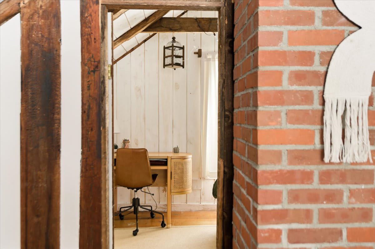 A glimpse through a doorway reveals a dedicated office space featuring a modern wooden desk with a cane front, a brown leather office chair, and white shiplap walls under a vaulted ceiling with exposed beams.