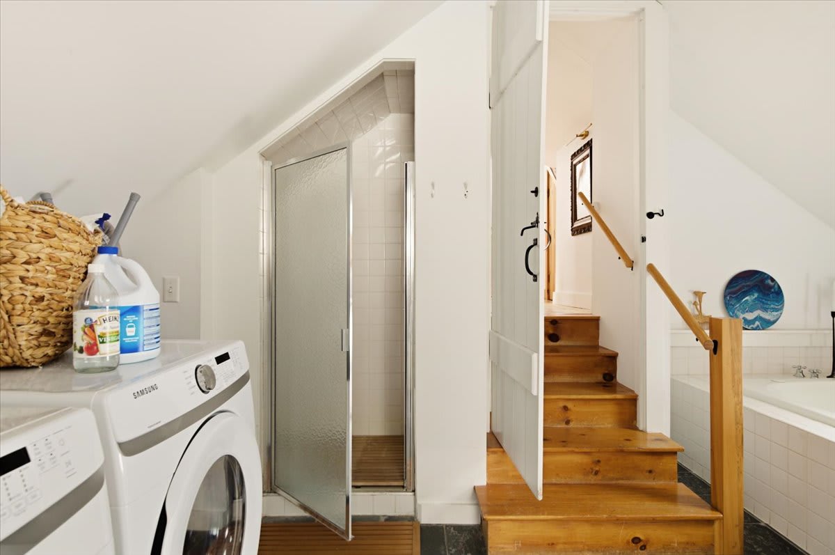 A compact shower stall with a glass door is visible next to a washing machine, with wooden stairs leading up. The space features white walls and dark floor tiles, indicating a combined bathroom and laundry area.