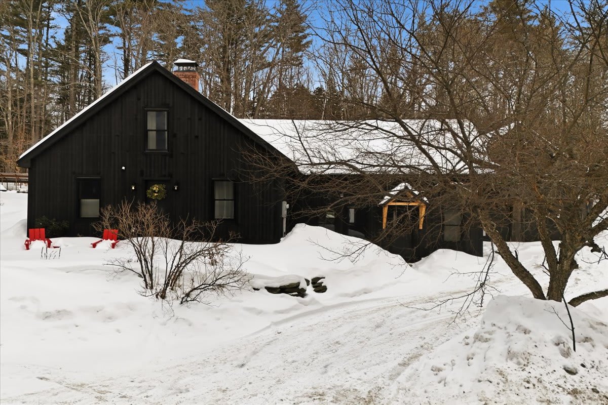 The black farmhouse-style house is nestled in a snowy landscape with bare trees, featuring a gabled roof and multiple windows. Red Adirondack chairs add a pop of color to the winter scene, with a clear driveway leading to the entrance.