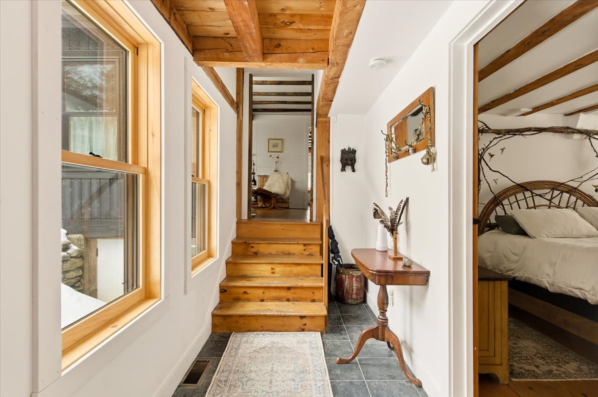 A bright entryway or hallway with slate tile flooring transitioning to wooden stairs leading to an upper level. A small wooden console table with decor and a mirror are visible, along with a glimpse into an adjacent bedroom.
