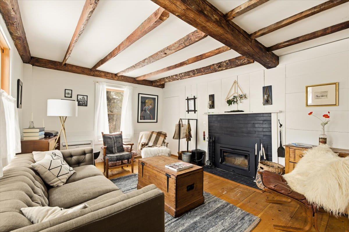 A cozy living room featuring a tufted sofa, a rustic wooden coffee table, and a black brick fireplace with a wood-burning insert. Exposed wooden ceiling beams and hardwood floors add to the farmhouse aesthetic.