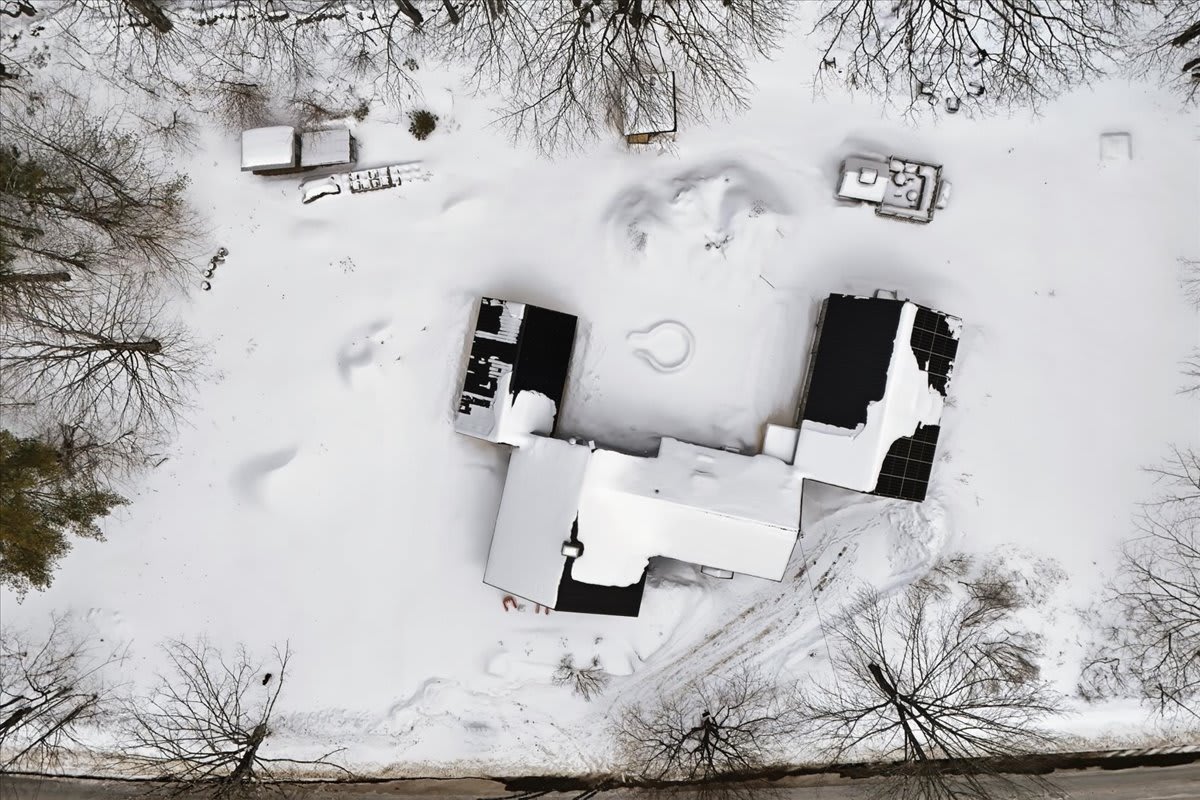 Overhead aerial view of the property, revealing a cluster of dark-roofed buildings partially covered in snow, nestled within a vast, snow-blanketed landscape dotted with bare trees.