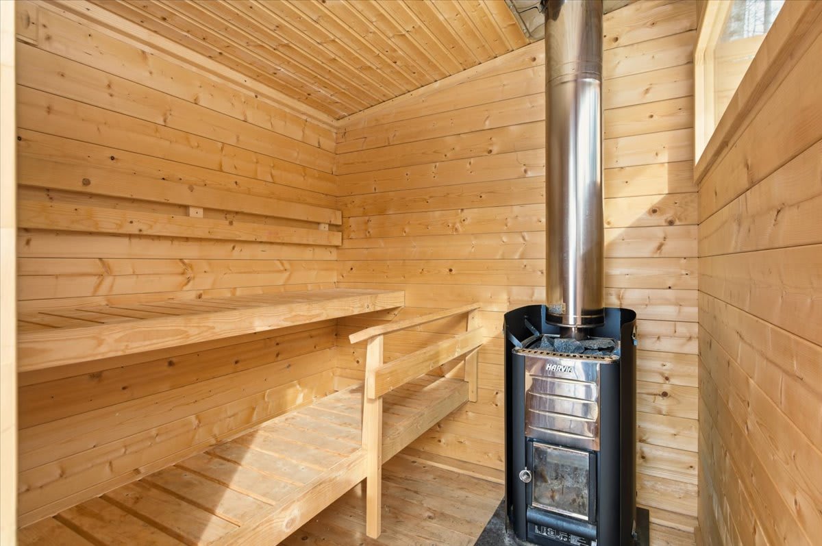 Interior of a cozy wooden sauna, featuring multi-level wooden benches, a prominent metal wood-burning stove with a chimney, and warm natural light.