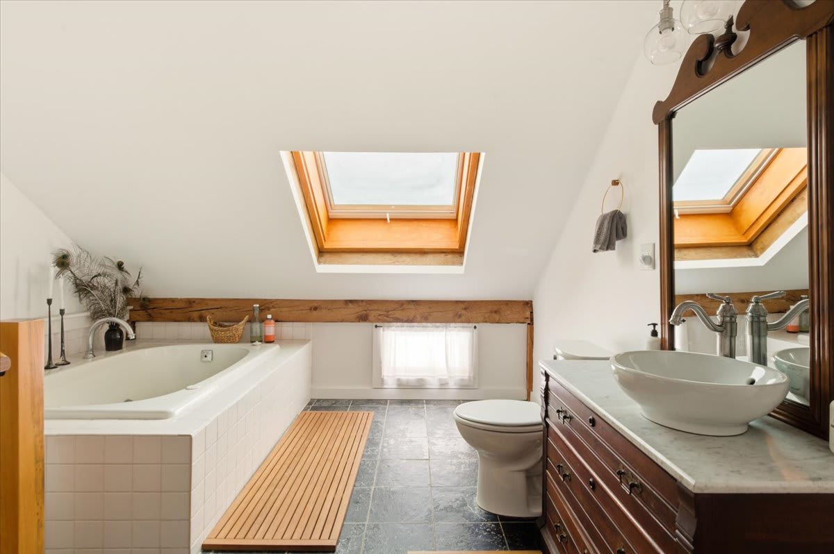 A bright attic bathroom with a white tiled bathtub, toilet, and a dark wood vanity with a vessel sink, all illuminated by a large skylight. Dark slate floor tiles contrast with white walls and a rustic exposed beam.