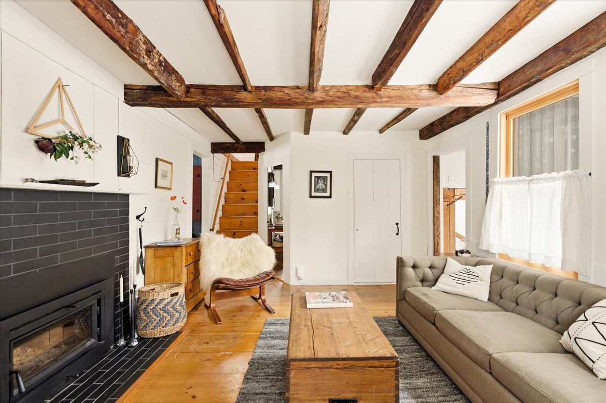 Wide view of a rustic living room with a tufted sofa, wooden coffee table, and a black brick fireplace. Exposed ceiling beams, hardwood floors, and a visible staircase leading upstairs enhance the farmhouse charm.