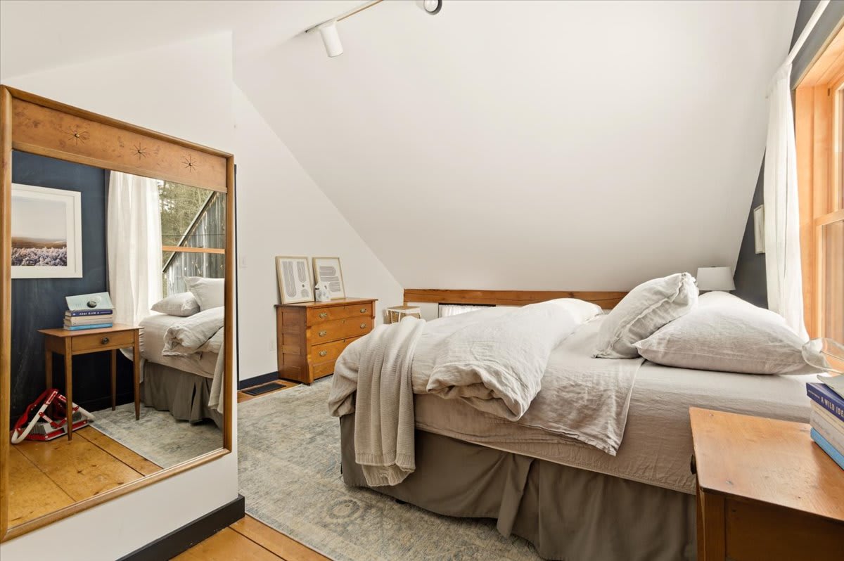 An attic bedroom with a full-length wooden mirror reflecting the room, a large bed, and a wooden side table. The room features sloped white ceilings, light hardwood floors, and a subtle area rug.