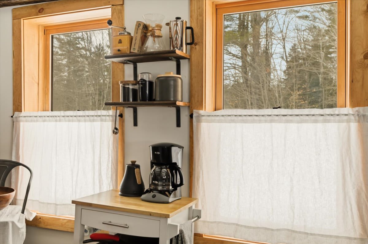 A bright kitchen corner with two large windows offering views of the snowy outdoors, adorned with white cafe curtains. A coffee station with a coffee maker and kettle sits on a small cart, beneath open shelving.