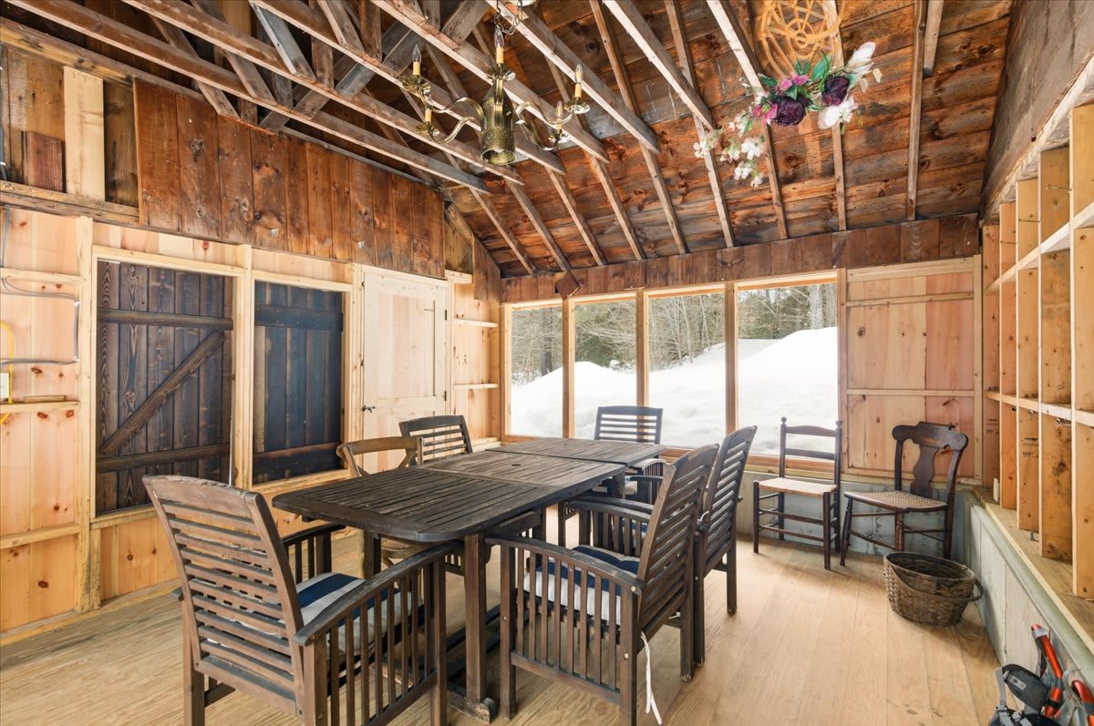 A rustic enclosed porch or sunroom features natural wood paneling on walls and ceiling, a large dark wooden dining table with slatted chairs, and expansive windows offering views of a snowy outdoor landscape.