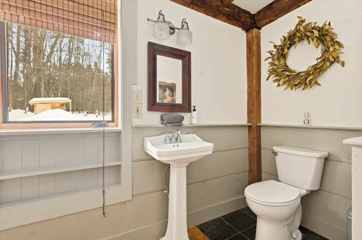 A rustic bathroom with a white pedestal sink, toilet, and light gray wainscoting. Exposed wooden beams and a window with a woven blind offer views of a snowy outdoor scene.
