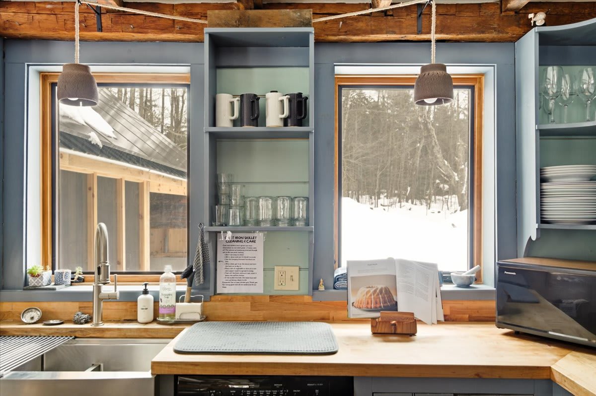 A bright kitchen area with light blue cabinetry, butcher block countertops, and a stainless steel sink. Two large windows overlook a snowy landscape, framed by exposed wooden beams.