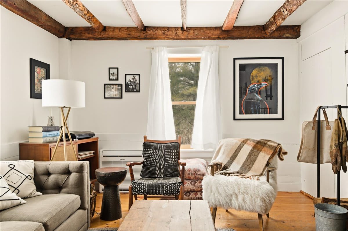 Bright living room with a tufted sofa, various armchairs, and a large window with sheer curtains. Exposed wooden ceiling beams, hardwood floors, and eclectic artwork contribute to a comfortable, lived-in feel.