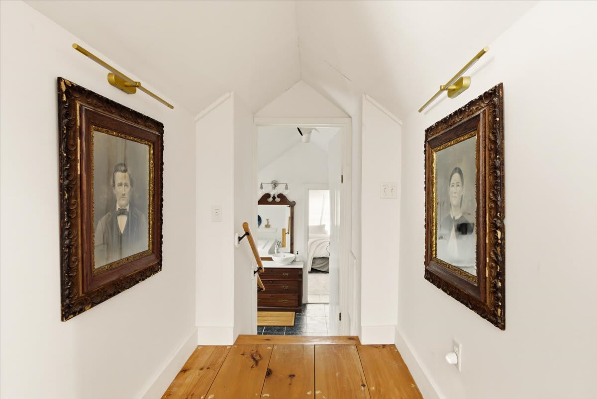 A narrow hallway with light hardwood floors features white walls adorned with two large, ornate framed portraits, leading towards a visible sink and mirror in a distant room.