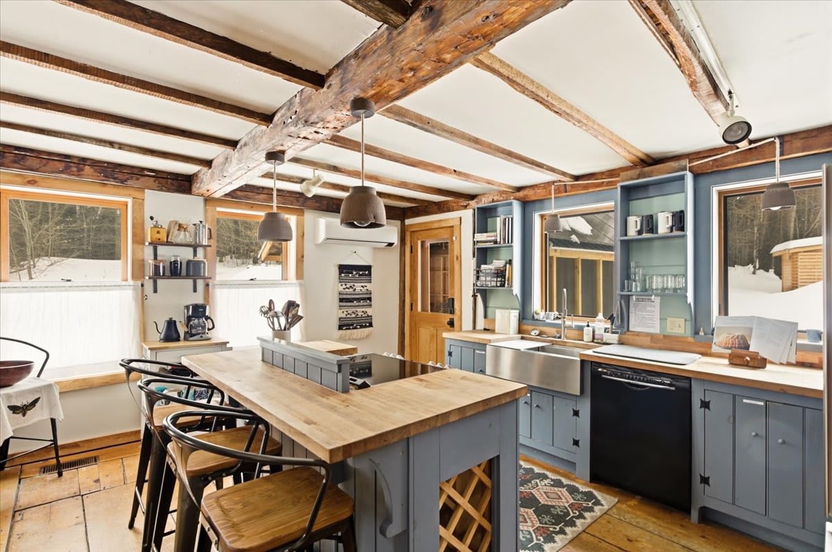 An open-concept kitchen featuring a large island with seating and a built-in stovetop, complemented by light blue cabinetry and butcher block countertops. Exposed wooden beams create a rustic yet functional space.