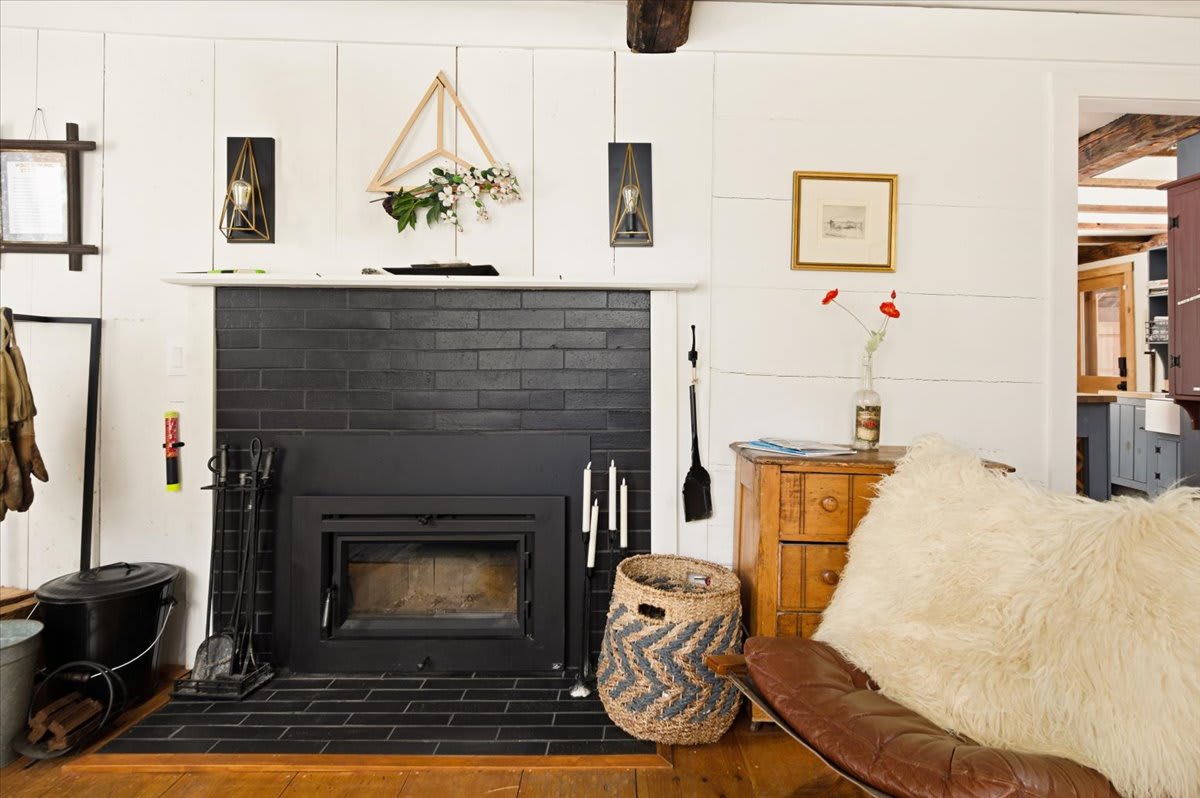 Close-up of a black brick fireplace with a wood-burning insert, flanked by modern sconces and a rustic wooden chest. The mantelpiece is adorned with simple decor, and a woven basket sits on the black tiled hearth.