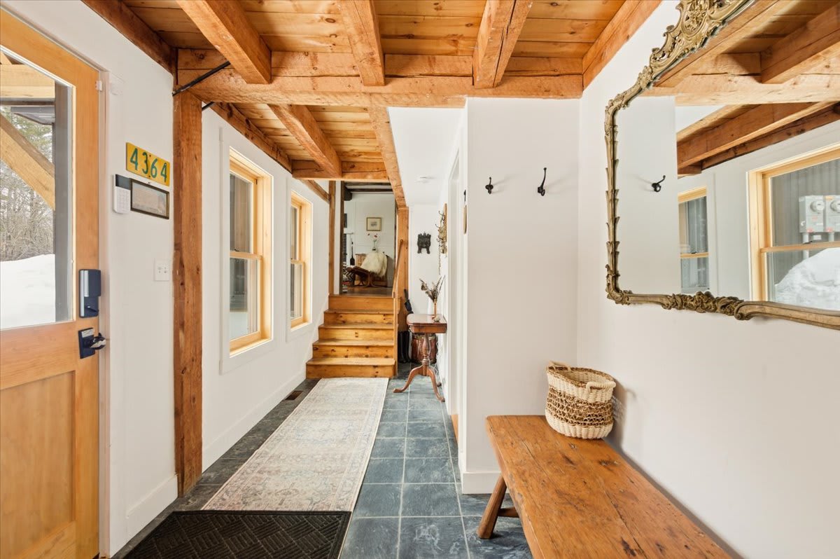 Long entryway featuring dark slate tile floors, exposed wooden ceiling beams, a rustic wooden bench, and a large ornate mirror. A wooden staircase is visible in the background, leading to another level.