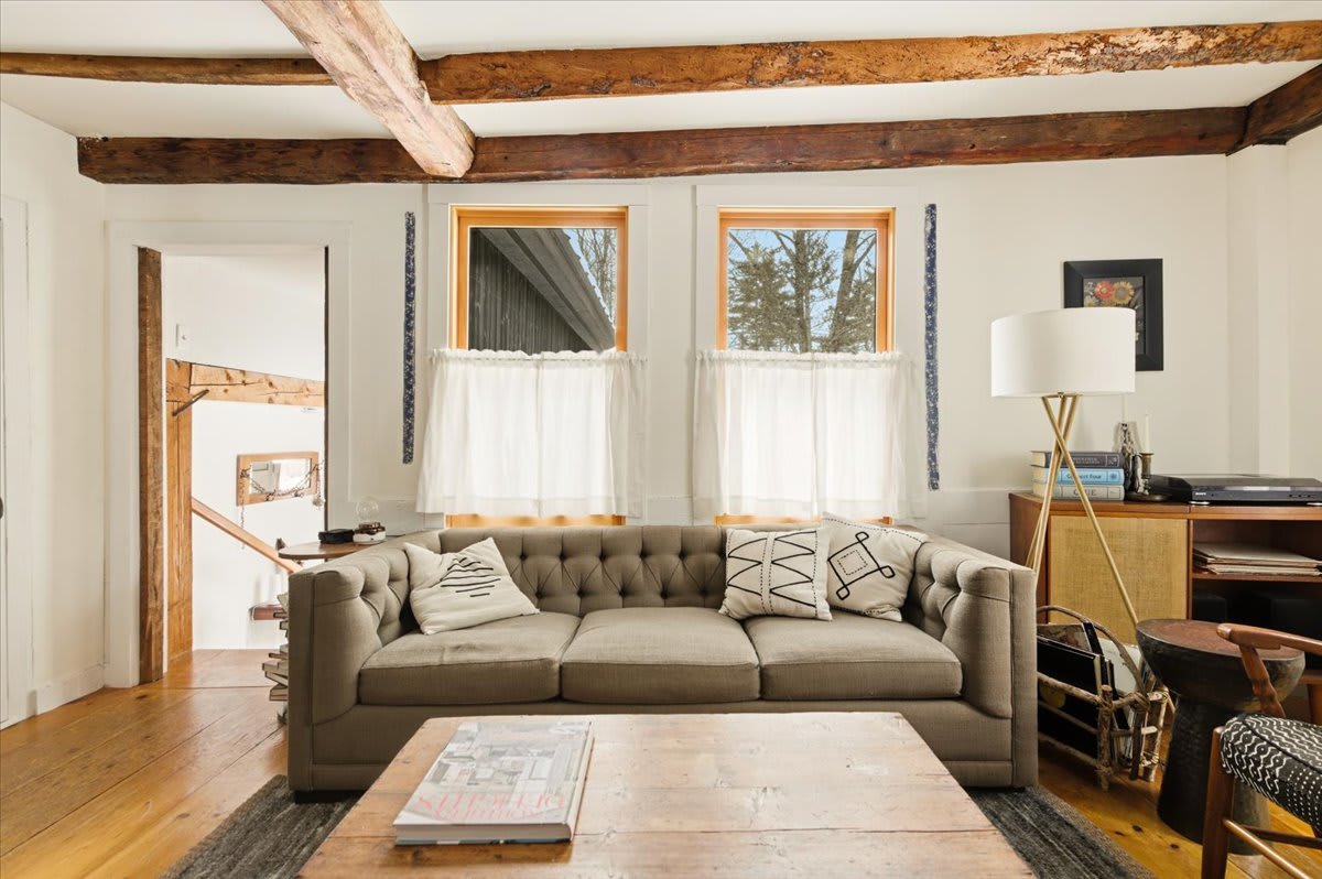 Bright living room featuring a tufted grey sofa, light wooden floors, exposed wooden ceiling beams, and two windows with sheer white curtains. A wooden coffee table and a modern floor lamp complete the space.