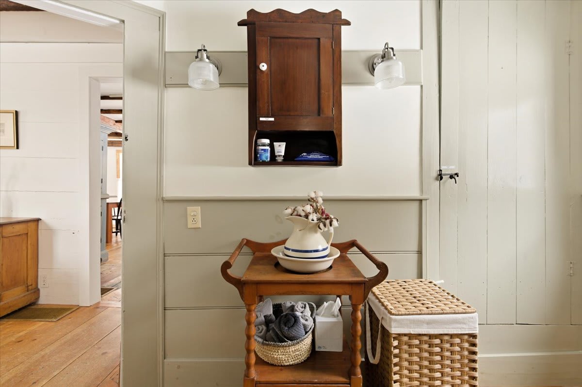 A transitional space adjacent to the bathroom, featuring a wooden serving cart with a decorative pitcher and a woven laundry basket. A dark wood wall cabinet with sconces hangs above light gray wainscoting.